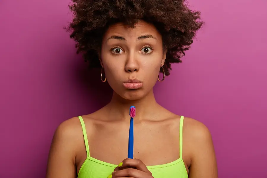 Oral hygiene concept. Disappointed dark skinned young woman looks sadly , purses lower lip, holds tooth brush, cleans teeth in morning, hears advice about dental care, poses indoor.